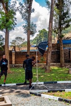 In a park or outdoor area, a person is holding a camera while using a large photography umbrella. Another individual stands nearby observing. Tall trees with green leaves and a blue sky with scattered clouds are visible in the background. The ground is covered with grass and a stone pathway.