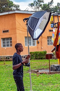 A person is holding a camera and a lighting setup outdoors. The background features an orange building with windows, greenery, and some colorful objects hanging.