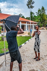 A photographer captures a woman posing outdoors in front of a modern building with red roofing and columns. The setting includes a large softbox for professional lighting. The scene is surrounded by greenery and trees, with a cloudy sky overhead.