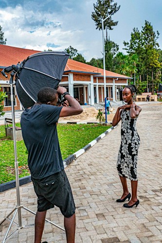 A photographer captures a woman posing outdoors in front of a modern building with red roofing and columns. The setting includes a large softbox for professional lighting. The scene is surrounded by greenery and trees, with a cloudy sky overhead.