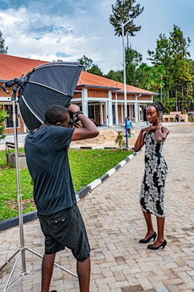 A photographer captures a woman posing outdoors in front of a modern building with red roofing and columns. The setting includes a large softbox for professional lighting. The scene is surrounded by greenery and trees, with a cloudy sky overhead.