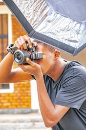A person is holding a camera up to their eye, ready to take a photograph. The individual is positioned under a large reflective photographic umbrella, likely for lighting purposes. The setting appears to be outdoors, with a blurred background of a building.