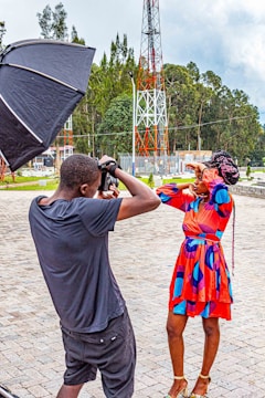 A photographer is taking a picture of a woman who is posing with one hand on her forehead, outside in an open area with a large telecom tower in the background. The woman is wearing a colorful, patterned dress and gold high-heeled shoes. There is a large, black reflective umbrella to the left, presumably used for lighting.