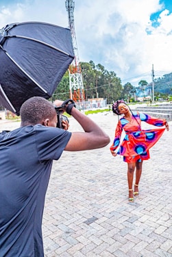 A photographer adjusts a large softbox while capturing a model posing in a vibrant, colorful dress. The setting is outdoors with a backdrop of greenery and a communication tower. The model appears to be mid-motion, showcasing the dress.