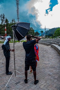 A photo shoot is taking place outdoors with a photographer holding a camera and capturing an image of a person in a bright, colorful outfit. A large softbox light is set up next to the photographer, and another person, possibly an assistant, stands nearby. The setting is a paved outdoor area surrounded by greenery, with a telecommunication tower and dramatic clouds in the background.