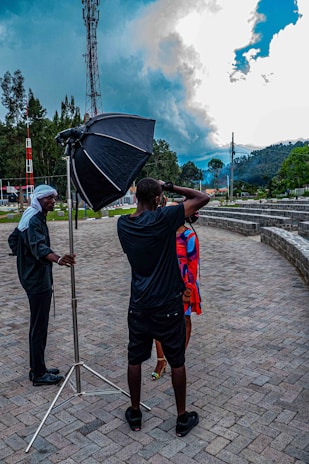 A photo shoot is taking place outdoors with a photographer holding a camera and capturing an image of a person in a bright, colorful outfit. A large softbox light is set up next to the photographer, and another person, possibly an assistant, stands nearby. The setting is a paved outdoor area surrounded by greenery, with a telecommunication tower and dramatic clouds in the background.