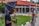 A photographer adjusts his camera while a woman stands nearby, in front of a building with a red roof and brick walls. The setting appears to be a formal outdoor location with a garden and pathways.