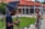 A photographer adjusts his camera while a woman stands nearby, in front of a building with a red roof and brick walls. The setting appears to be a formal outdoor location with a garden and pathways.