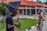 A photographer adjusts his camera while a woman stands nearby, in front of a building with a red roof and brick walls. The setting appears to be a formal outdoor location with a garden and pathways.