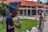 A photographer adjusts his camera while a woman stands nearby, in front of a building with a red roof and brick walls. The setting appears to be a formal outdoor location with a garden and pathways.