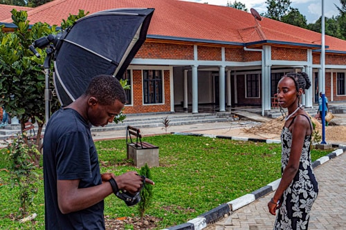 A photographer adjusts his camera while a woman stands nearby, in front of a building with a red roof and brick walls. The setting appears to be a formal outdoor location with a garden and pathways.