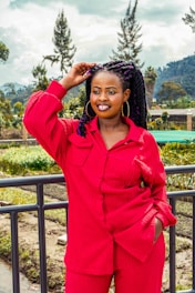 A person with long braided hair is smiling and standing outdoors near a railing with a backdrop of trees and distant hills. The individual is wearing vibrant red attire and hoop earrings, and they are touching their hair with one hand.