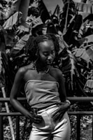 A woman stands confidently against a railing, with a backdrop of large leafy foliage. She wears a strapless top and light pants, exuding a poised stance. The image is black and white, creating a dramatic contrast between the subject and the lush surroundings.