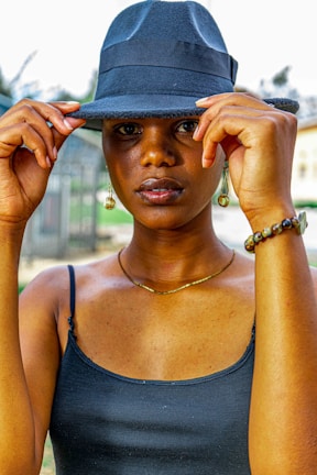 A person wearing a dark blue fedora hat and a matching tank top, with their hands adjusting the hat. They are accessorized with dangling earrings, a bracelet, and a necklace. The background is slightly blurred, suggesting an outdoor setting.