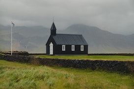 A small, black wooden church stands in a grassy area surrounded by a low stone wall, with misty mountains in the background. A flagpole with the Icelandic flag rises beside the church, under cloudy skies.