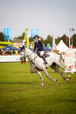 A person wearing a riding outfit, including a helmet, is riding a white horse at an equestrian event. The horse is in motion, suggesting a competitive setting. In the background, there are various tents and banners, indicating an outdoor event with spectators.