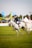 A person wearing a riding outfit, including a helmet, is riding a white horse at an equestrian event. The horse is in motion, suggesting a competitive setting. In the background, there are various tents and banners, indicating an outdoor event with spectators.