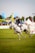 A person wearing a riding outfit, including a helmet, is riding a white horse at an equestrian event. The horse is in motion, suggesting a competitive setting. In the background, there are various tents and banners, indicating an outdoor event with spectators.