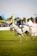 A person wearing a riding outfit, including a helmet, is riding a white horse at an equestrian event. The horse is in motion, suggesting a competitive setting. In the background, there are various tents and banners, indicating an outdoor event with spectators.