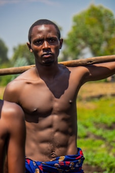 A shirtless man with defined muscles stands outdoors. He carries a wooden pole over his shoulder. The background shows greenery and a blurred natural environment.