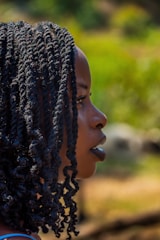 A side profile of a person with braided hair, set against a blurred, natural background with green and earthy tones.