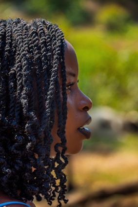 A side profile of a person with braided hair, set against a blurred, natural background with green and earthy tones.