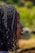 A side profile of a person with braided hair, set against a blurred, natural background with green and earthy tones.