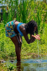 A person is bending over to scoop water from a clear stream with their hands. They are wearing a blue top and colorful patterned shorts. Surrounding the scene are tall, green grasses and vibrant foliage, suggesting a lush, natural environment.