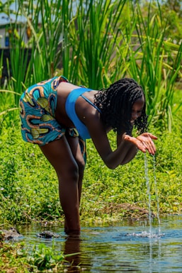 A person is bending over to scoop water from a clear stream with their hands. They are wearing a blue top and colorful patterned shorts. Surrounding the scene are tall, green grasses and vibrant foliage, suggesting a lush, natural environment.