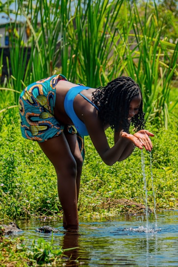 A person is bending over to scoop water from a clear stream with their hands. They are wearing a blue top and colorful patterned shorts. Surrounding the scene are tall, green grasses and vibrant foliage, suggesting a lush, natural environment.