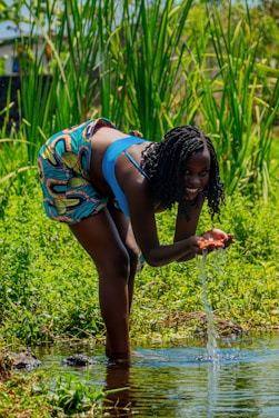 A woman in colorful attire is gathering water from a small stream or pond, surrounded by lush green plants. She appears to be smiling and engaging in a playful manner. The environment is vibrant, with tall reeds and bright sunlight.