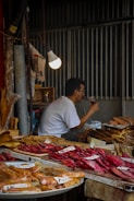 A market stall displays a variety of dried and smoked fish on trays, with handwritten labels. A man in a white shirt is seated, drinking from a clear glass under a glowing light bulb. The background features dimly lit corrugated metal walls.