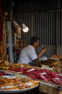 A market stall displays a variety of dried and smoked fish on trays, with handwritten labels. A man in a white shirt is seated, drinking from a clear glass under a glowing light bulb. The background features dimly lit corrugated metal walls.