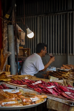 A market stall displays a variety of dried and smoked fish on trays, with handwritten labels. A man in a white shirt is seated, drinking from a clear glass under a glowing light bulb. The background features dimly lit corrugated metal walls.