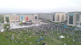 A large crowd of people is gathered on a green lawn and in front of a modern building complex with large arches and decorative panels. The scene is outdoors with lush grass and the building is adorned with large national flags. People appear to be enjoying a public event or gathering, sitting and walking around the open area.