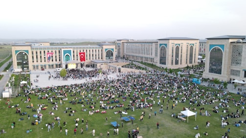 A large crowd of people is gathered on a green lawn and in front of a modern building complex with large arches and decorative panels. The scene is outdoors with lush grass and the building is adorned with large national flags. People appear to be enjoying a public event or gathering, sitting and walking around the open area.