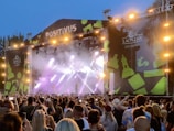 A large outdoor concert stage with bright lights and a vibrant backdrop is featured. The stage is labeled 'POSITIVUS' and is adorned with abstract green patterns. A crowd of people standing in front of the stage, many holding up phones to take photos or videos, indicating a lively atmosphere.