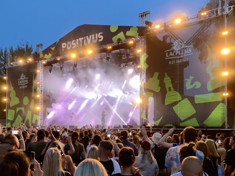 A large outdoor concert stage with bright lights and a vibrant backdrop is featured. The stage is labeled 'POSITIVUS' and is adorned with abstract green patterns. A crowd of people standing in front of the stage, many holding up phones to take photos or videos, indicating a lively atmosphere.