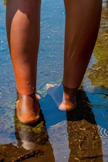 A person's bare feet are immersed in clear, shallow water with visible reflections on the surface. The sunlight illuminates the skin, creating a contrast with the dark water and green algae-covered stone beneath the feet.