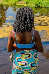 A person with intricately braided hair stands near the edge of a body of water, wearing a vibrant patterned wrap and a blue top. The water is partly covered with green vegetation, and there are patches of algae on the surface. The surroundings suggest a natural or outdoor setting with greenery.