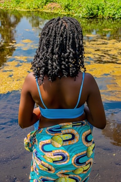 A person with intricately braided hair stands near the edge of a body of water, wearing a vibrant patterned wrap and a blue top. The water is partly covered with green vegetation, and there are patches of algae on the surface. The surroundings suggest a natural or outdoor setting with greenery.