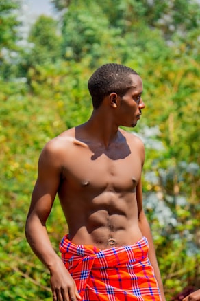 A young man with a toned physique stands outdoors, wrapped in a colorful, red and blue patterned cloth around his waist. The background is filled with lush green foliage, suggesting a natural setting.
