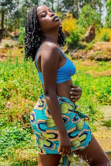 A person with braided hair stands outdoors in a vibrant, natural setting. They wear a bright blue top and a colorful patterned wrap skirt. The background is lush with greenery, possibly a field or garden, and the lighting suggests a sunny day.