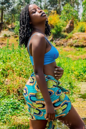 A person with braided hair stands outdoors in a vibrant, natural setting. They wear a bright blue top and a colorful patterned wrap skirt. The background is lush with greenery, possibly a field or garden, and the lighting suggests a sunny day.