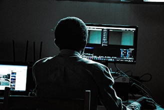 A person is sitting in front of two computer screens in a dimly lit room. One screen shows a video editing software interface, while the other displays a webpage, possibly YouTube. The person is in shadow, with the light from the screens illuminating parts of their silhouette. Various computer peripherals and cables are visible on the desk.