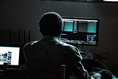 A person is sitting in front of two computer screens in a dimly lit room. One screen shows a video editing software interface, while the other displays a webpage, possibly YouTube. The person is in shadow, with the light from the screens illuminating parts of their silhouette. Various computer peripherals and cables are visible on the desk.
