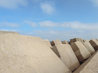 Large concrete blocks are stacked and spread out under a bright blue sky with white clouds. The structures appear to be part of a breakwater or sea defense system, providing a contrast to the expansive open sky above.