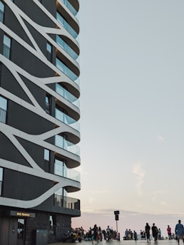 A modern multi-story building with a distinctive architectural design featuring curved white lines along its exterior. The building has several balconies with glass railings. To the right, there is a gathering of people and bicycles near the waterfront, with a vast, open sky in the background.