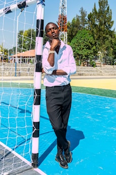 A person in formal attire stands leaning against a soccer goalpost on a vibrant blue sports court. The individual, wearing a white shirt and black trousers, appears contemplative. Surrounding the court are tall trees and a communication tower, under a clear blue sky.