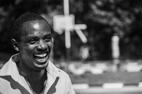 A black and white photograph depicts a person laughing, with a broad smile, captured mid-expression. The background is blurred, hinting at an outdoor setting with a basketball hoop visible.